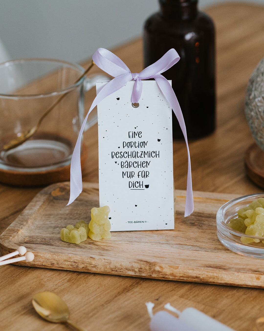 Teebären Beschützmich-Bärchen mit fliederfarbener Schleife auf Holzbrett – geschenkfertig verpackt.