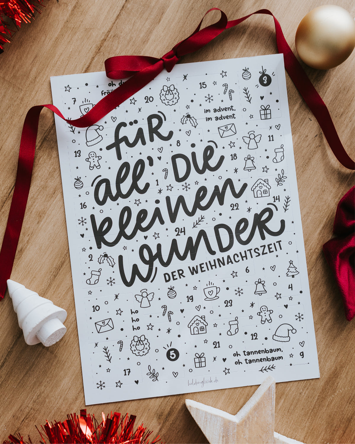 Adventskalender von oben auf Holztisch mit rotem Band und Weihnachtskugel.