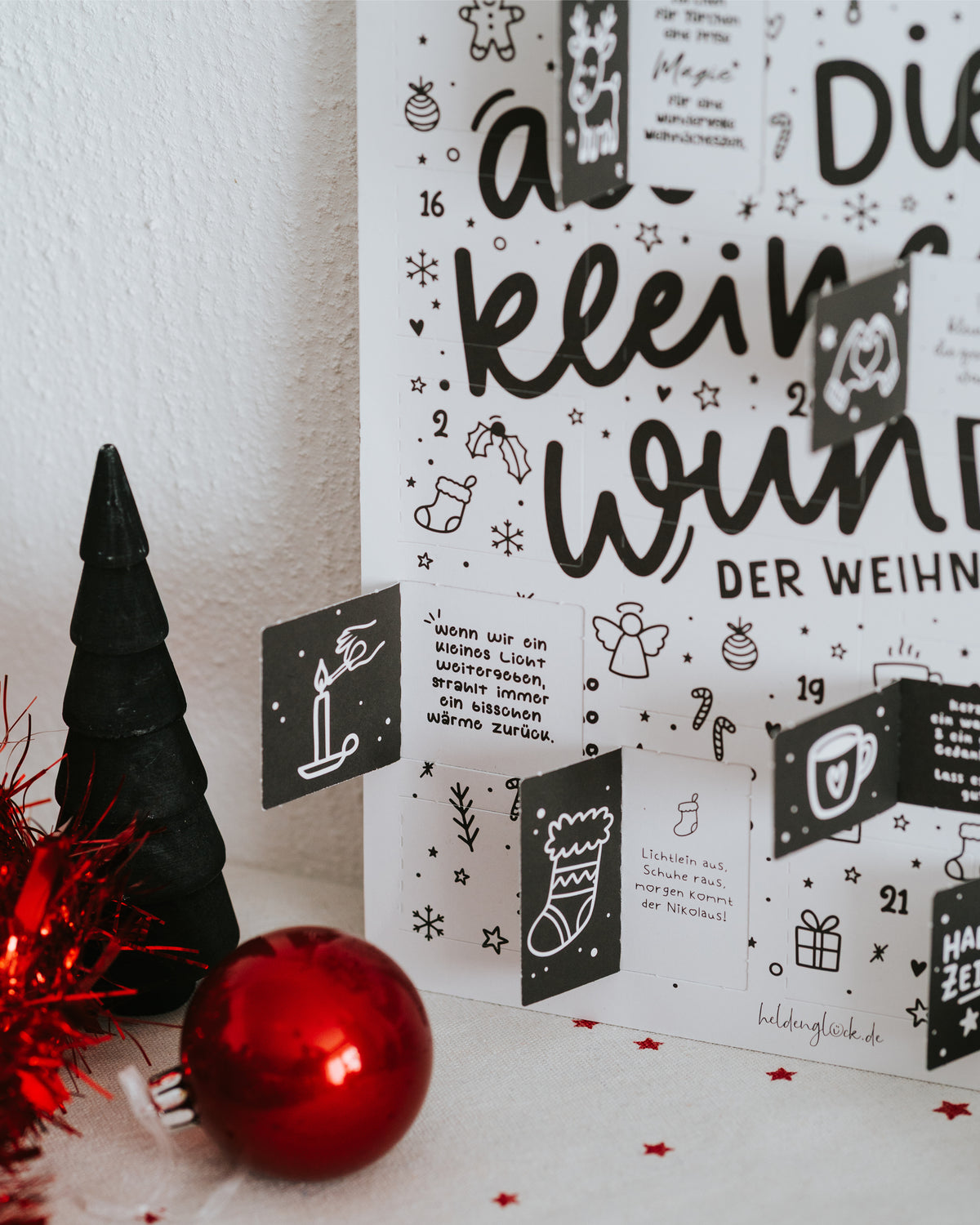 Geöffnete Türchen des Adventskalenders mit Illustrationen und Sprüchen, daneben rote Kugel und Deko-Tanne.