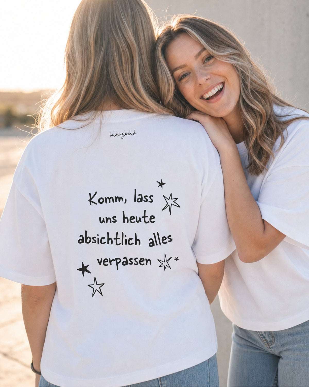 Zwei Frauen in weißen T-Shirts; auf einem Rücken steht „Komm, lass uns heute absichtlich alles verpassen“, umgeben von Sternen.