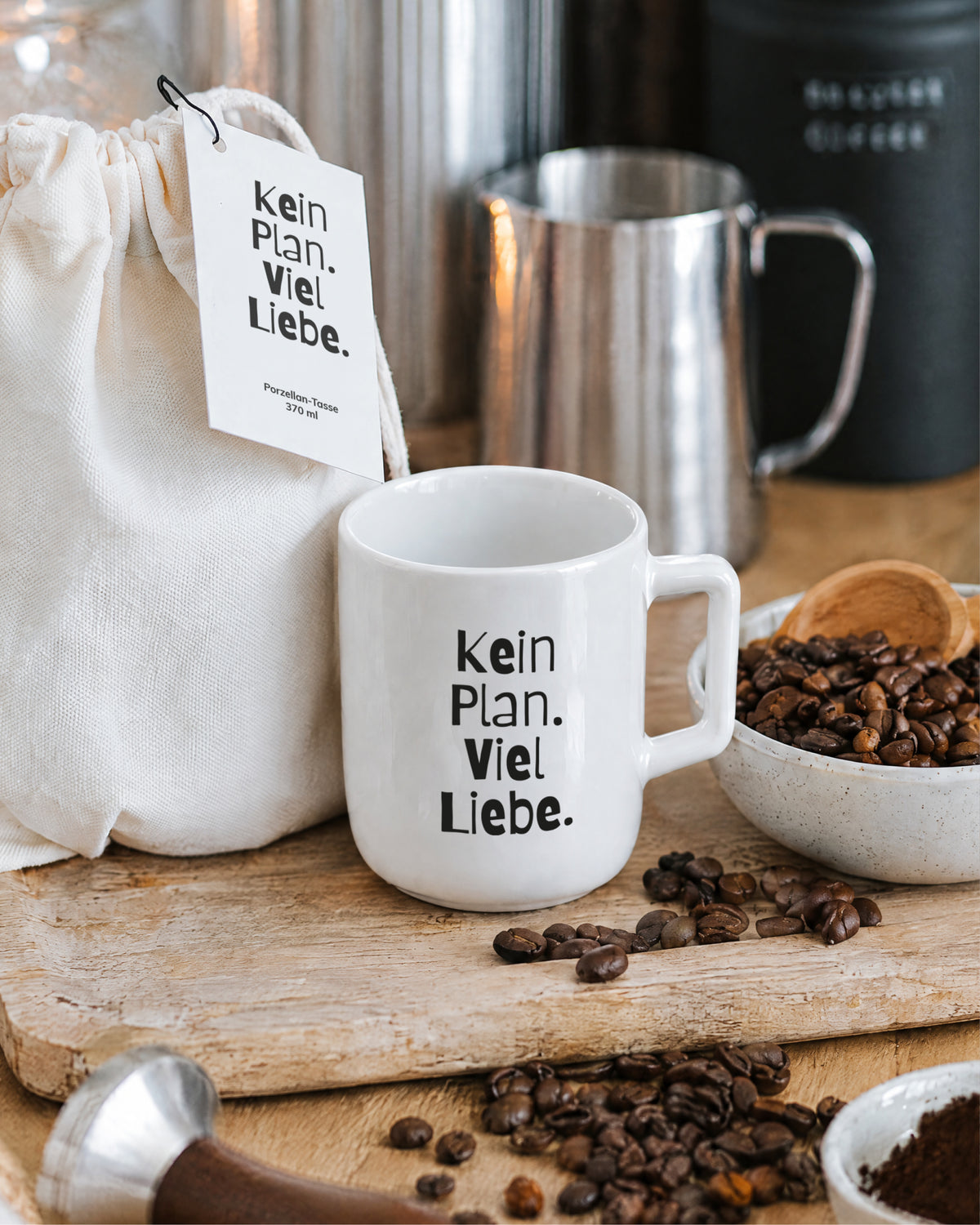 Porzellantasse „Kein Plan. Viel Liebe.“ auf Holzbrett, daneben Kaffeebohnen.