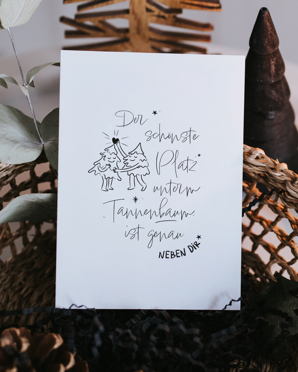 Weihnachtspostkarte „Der schönste Platz“ mit Tannenbaum-Illustration, aufgestellt zwischen Eukalyptuszweig und Holz-Christbäumen