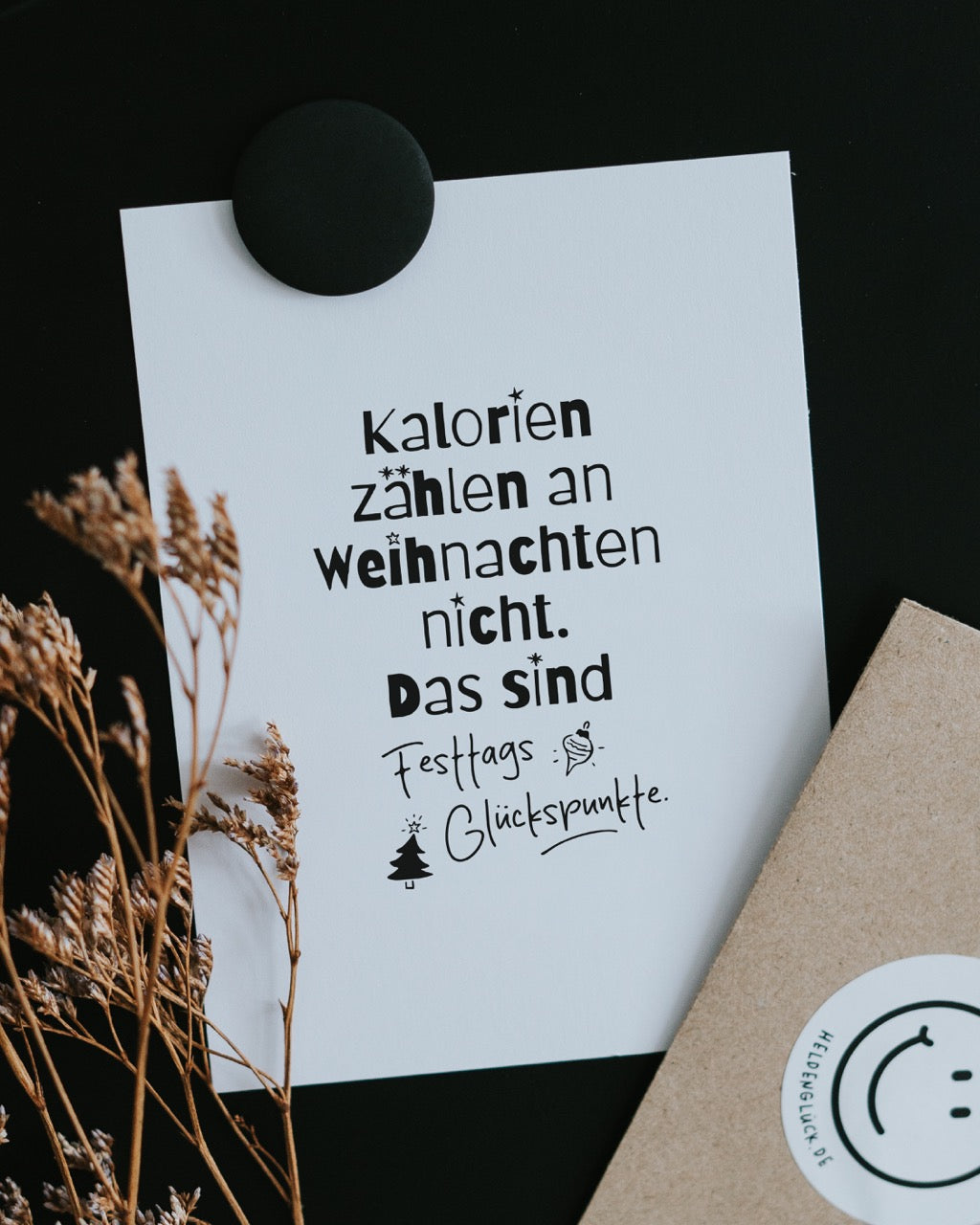Postkarte „Festtags-Glückspunkte“ mit Spruch zu Weihnachtskalorien, an einer schwarzen Pinnwand mit Magnet befestigt, daneben Trockengräser und ein Kraftpapier-Umschlag mit Heldenglück-Sticker