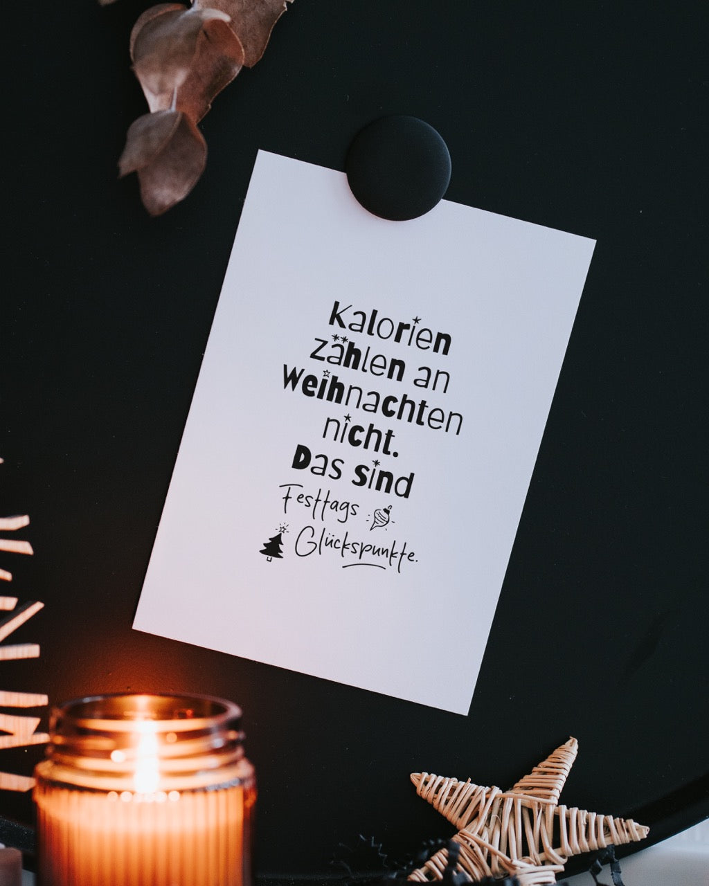Postkarte „Kalorien zählen an Weihnachten nicht“ an schwarzer Magnettafel, umrahmt von getrockneten Blättern, Kerzenlicht und einem Rattan-Stern