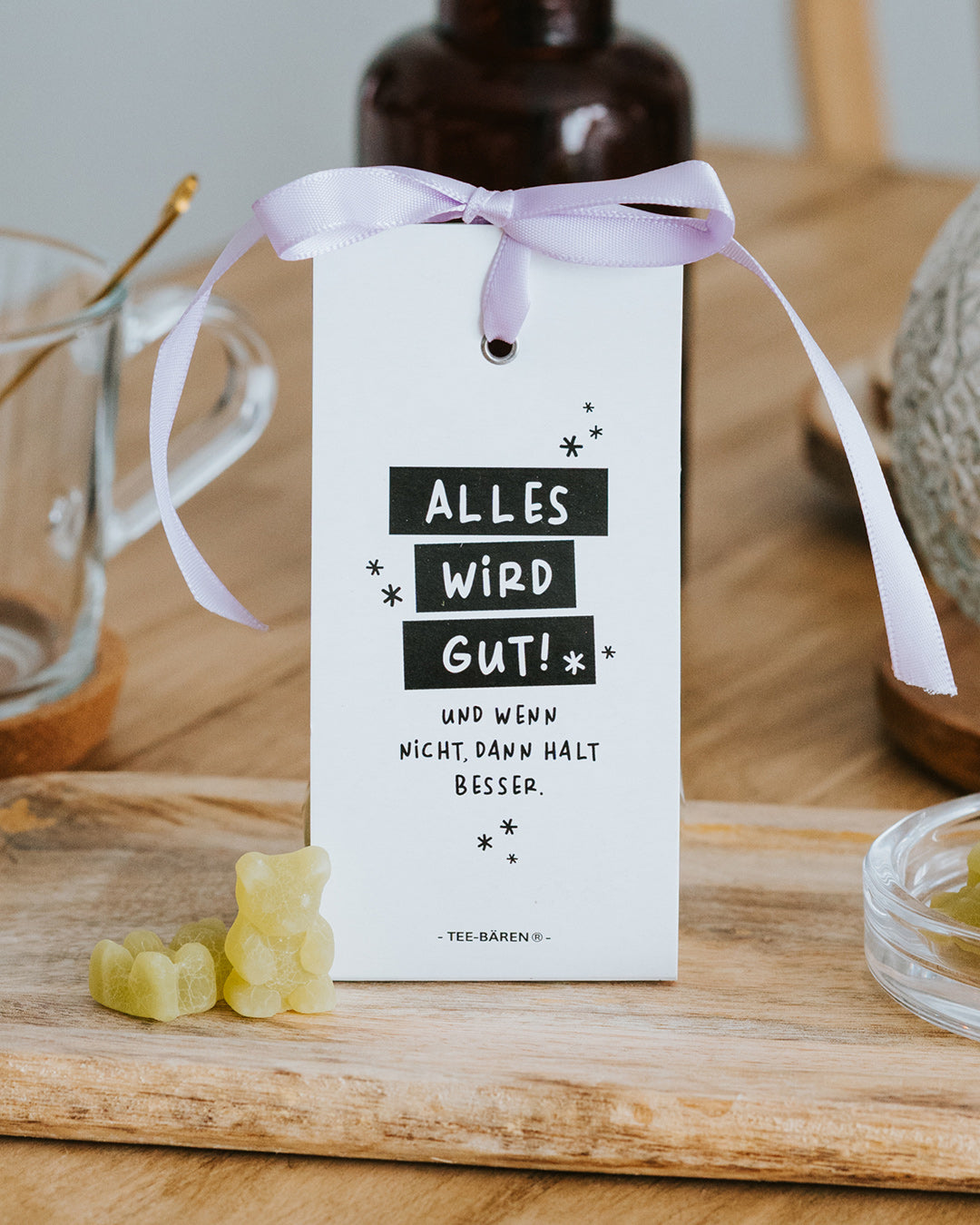Teebären Alles wird gut mit fliederfarbener Schleife auf Holzbrett – geschenkfertig verpackt.