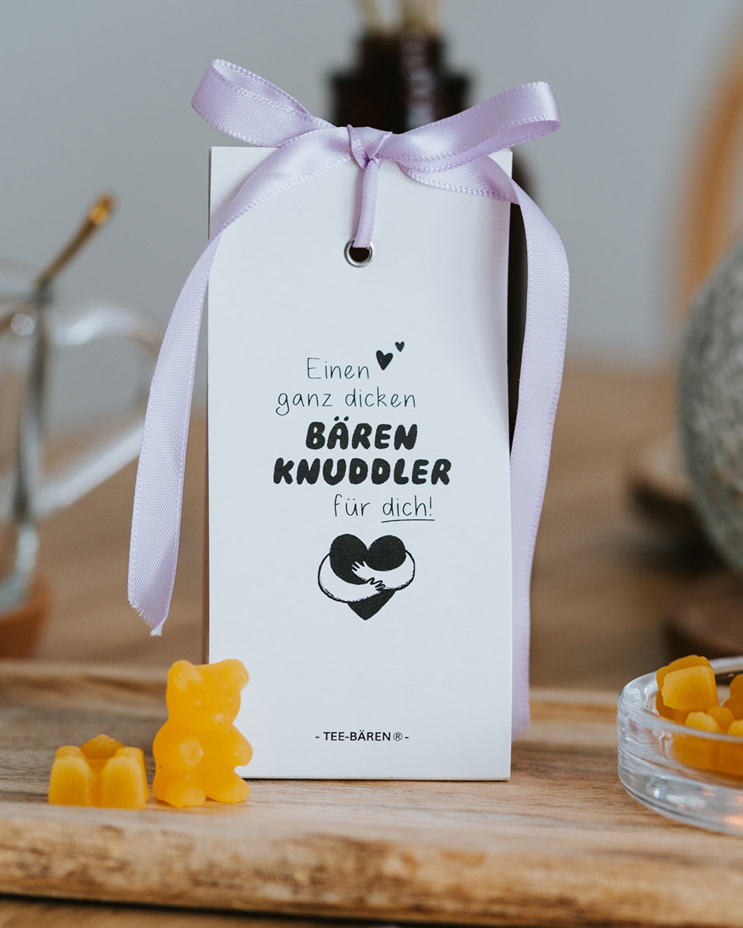 Teebären Bärenknuddler mit fliederfarbener Schleife auf Holzbrett – geschenkfertig verpackt, gelbe Bärchen davor.