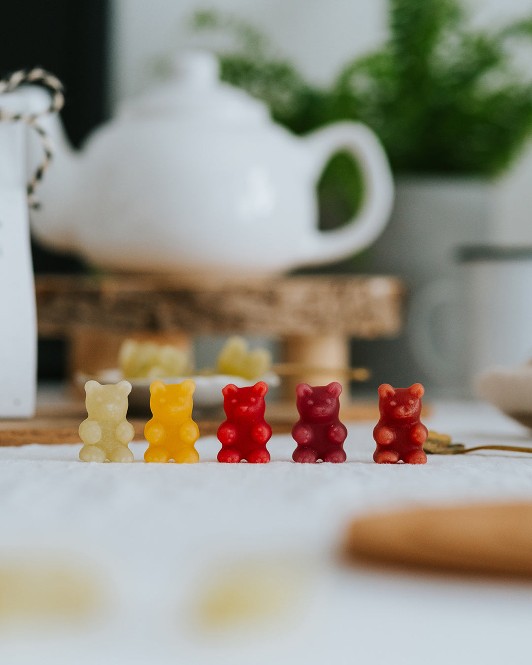 Fünf Fruchtgummi-Bärchen in allen Sorten in einer Reihe, dahinter eine weiße Teekanne.