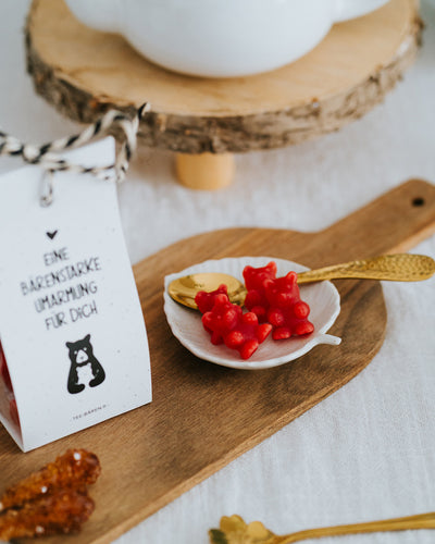 Rote Fruchtgummi-Bärchen im Schälchen mit Goldlöffel, Teebären-Tüte links angeschnitten.