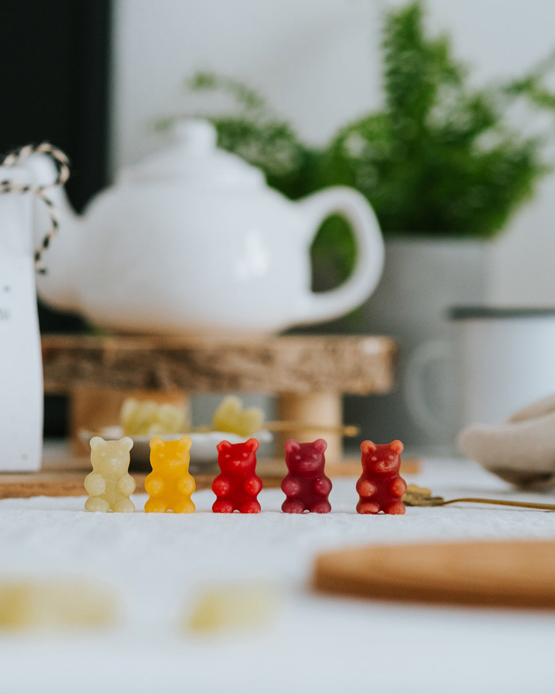 Fünf Fruchtgummi-Bärchen in allen Sorten in einer Reihe vor weißer Teekanne.