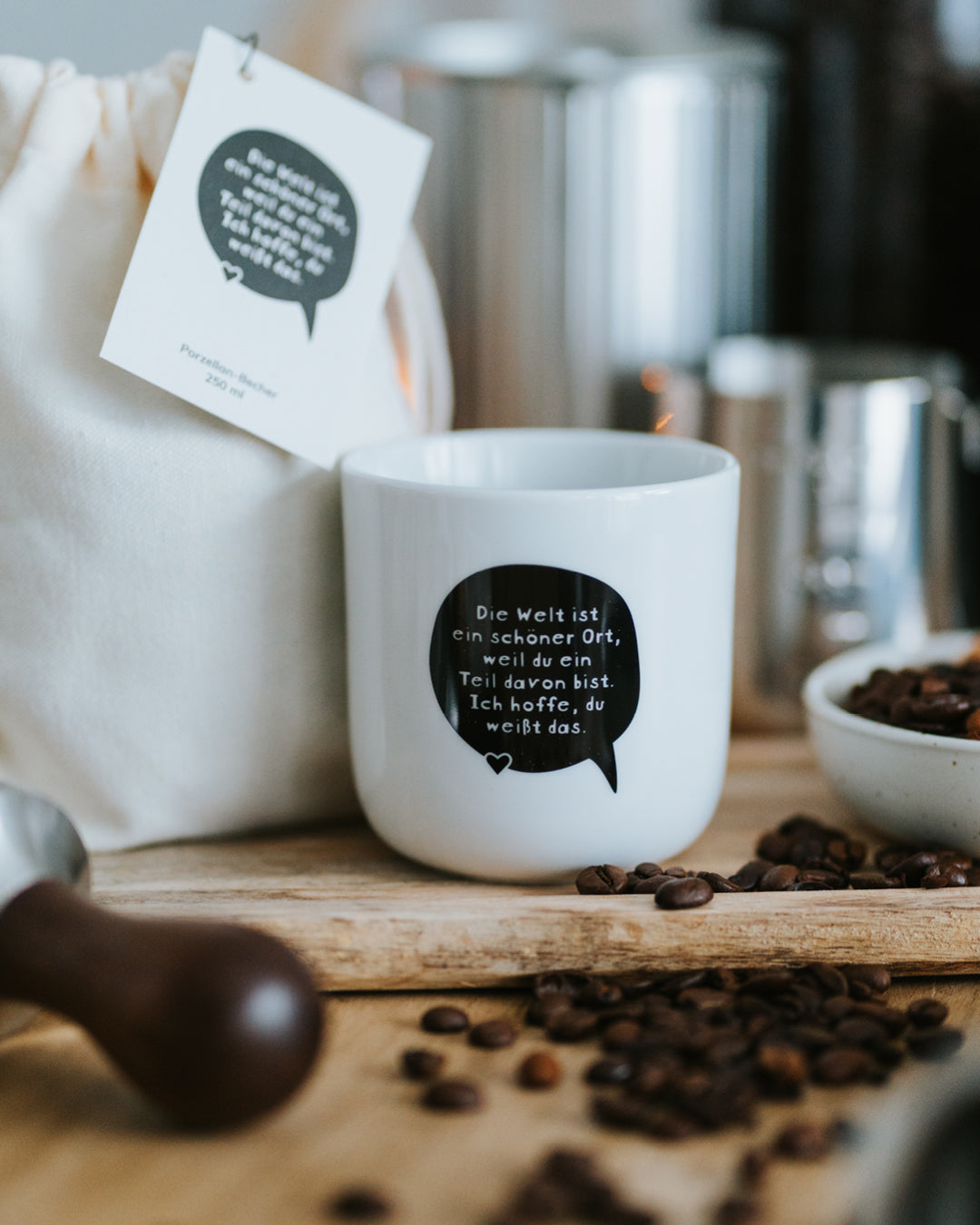 Porzellanbecher „Die Welt ist ein schöner Ort“ neben Baumwollbeutel, Tamper und Kaffeebohnen.