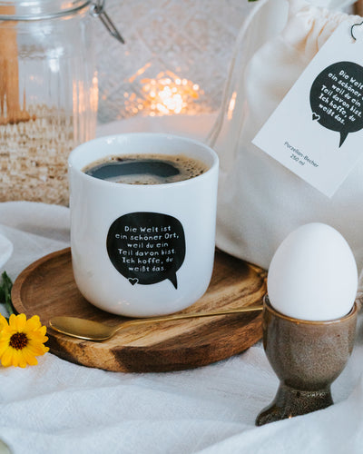 Porzellanbecher „Die Welt ist ein schöner Ort“ mit Kaffee auf gedecktem Frühstückstisch mit Ei.