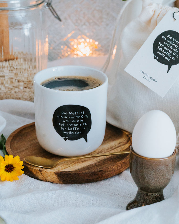 Porzellanbecher „Die Welt ist ein schöner Ort“ mit Kaffee auf gedecktem Frühstückstisch mit Ei.