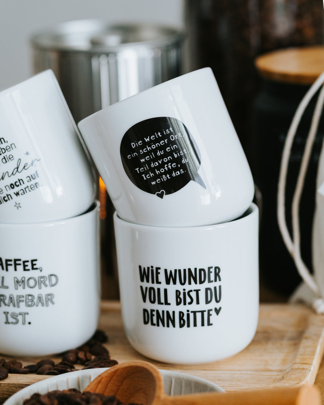 Gestapelte Heldenglück-Porzellanbecher, „Die Welt ist ein schöner Ort“ oben, dazu Kaffeebohnen.