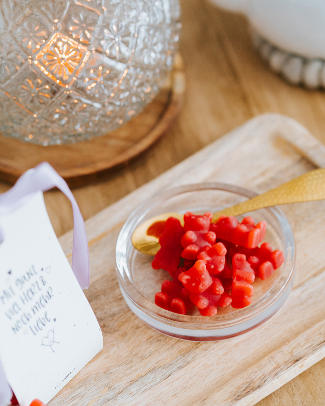 Rote Fruchtgummi-Bärchen in der Glasschale mit Goldlöffel vor Windlicht, Tüte links angeschnitten.