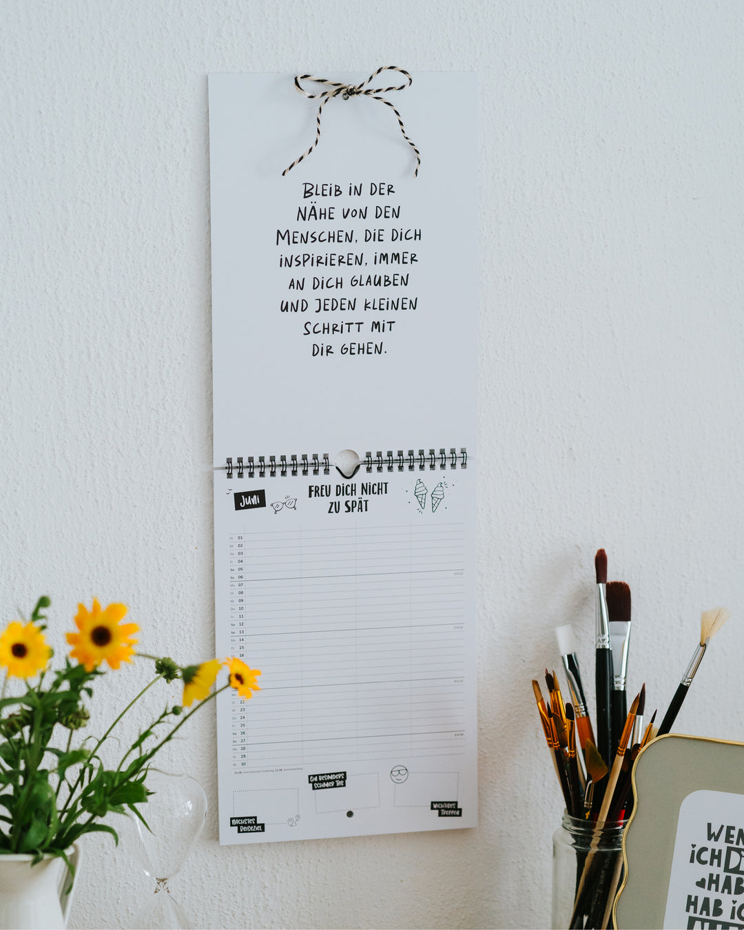 Familienkalender an der Wand mit aufgeschlagener Spruchseite – jeden Monat ein neuer Impuls, daneben Sonnenblumen und Pinsel.