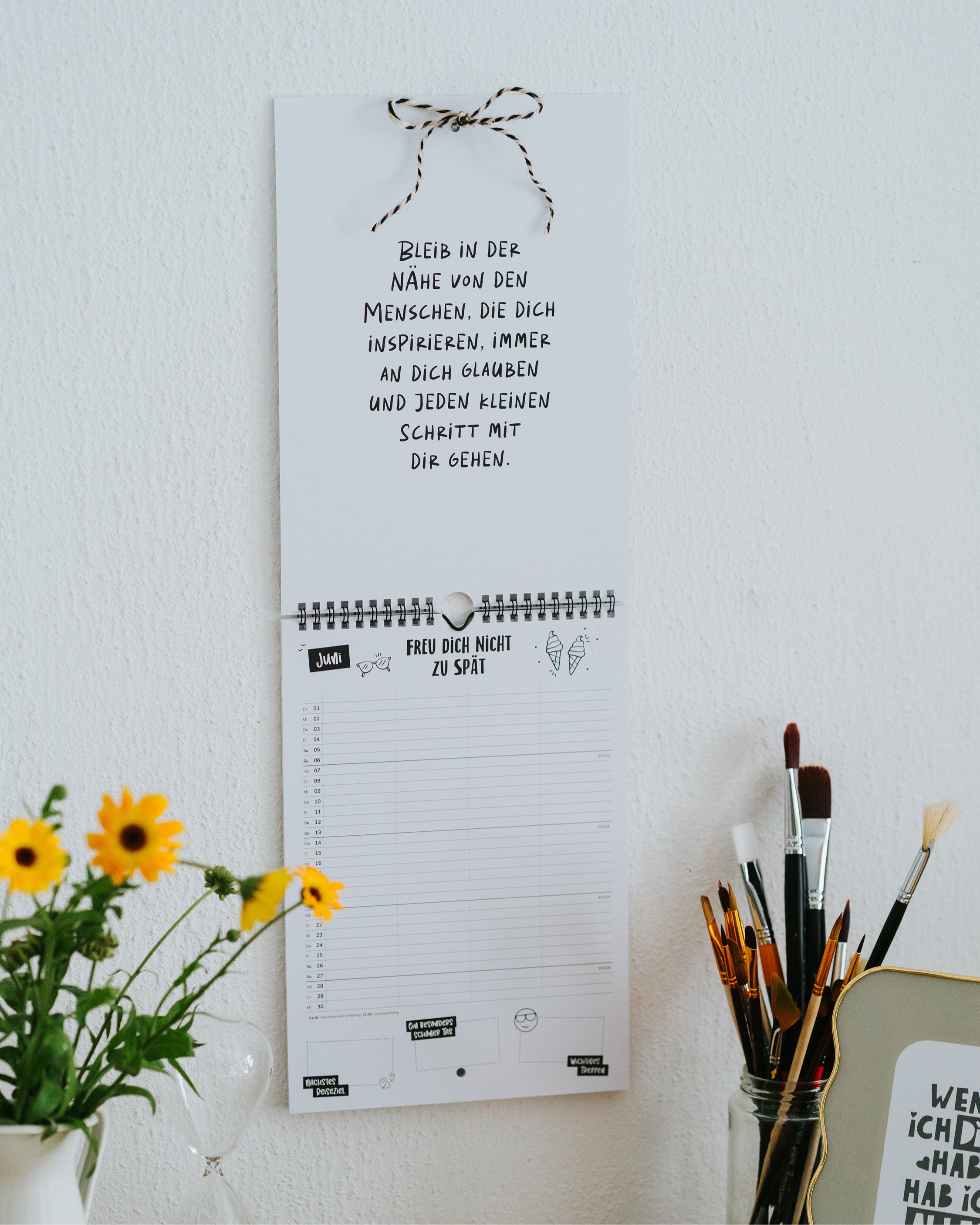 Familienkalender an der Wand mit aufgeschlagener Spruchseite – jeden Monat ein neuer Impuls, daneben Sonnenblumen und Pinsel.