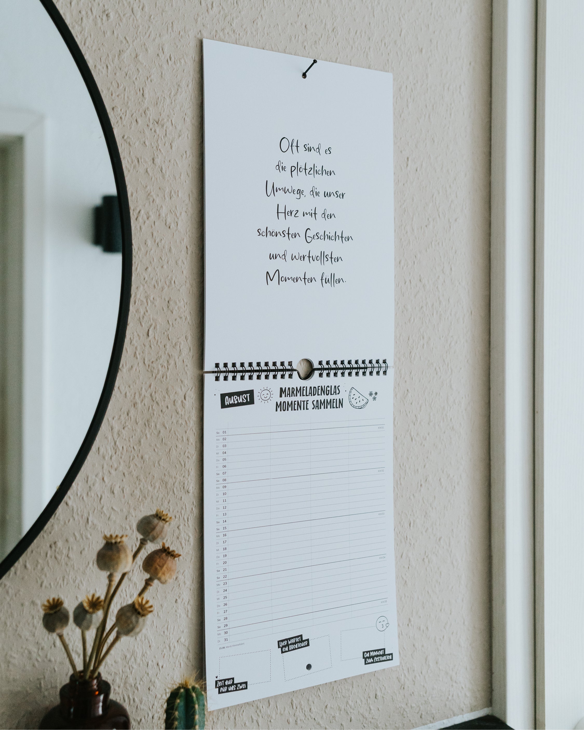 Familienkalender neben rundem Spiegel an der Wand – mit Sprüchen und Ferienübersicht, Trockenblumen und Kaktus daneben.