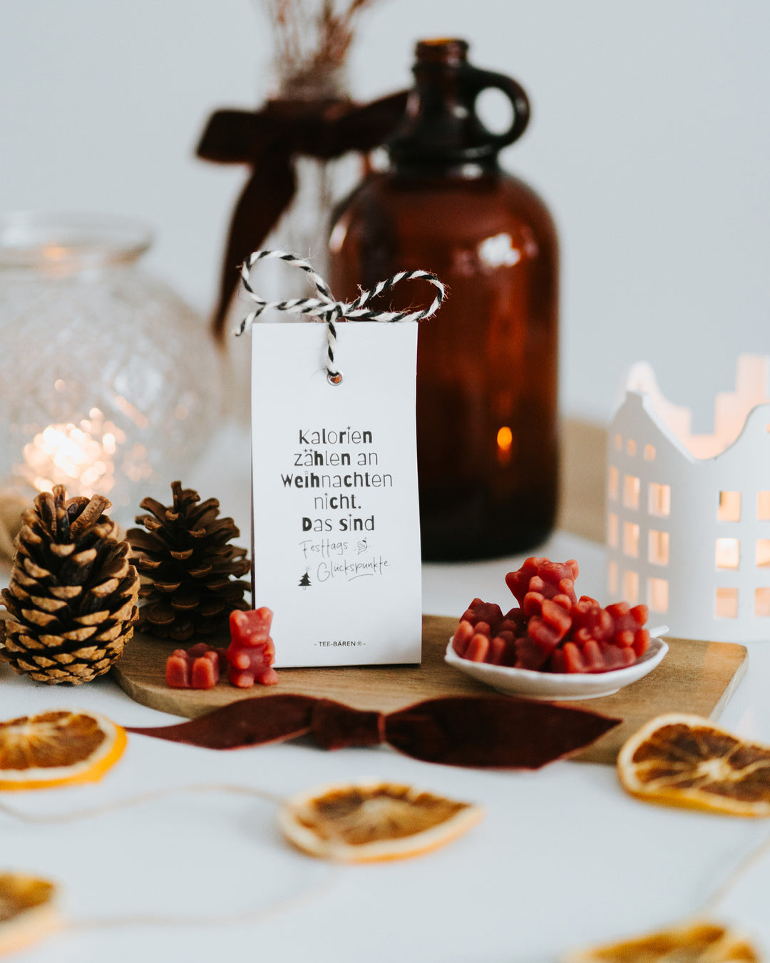 Teebären „Festtags-Glückspunkte“ auf weihnachtlich gedecktem Tisch mit Windlicht, Zapfen und Orangenscheiben.