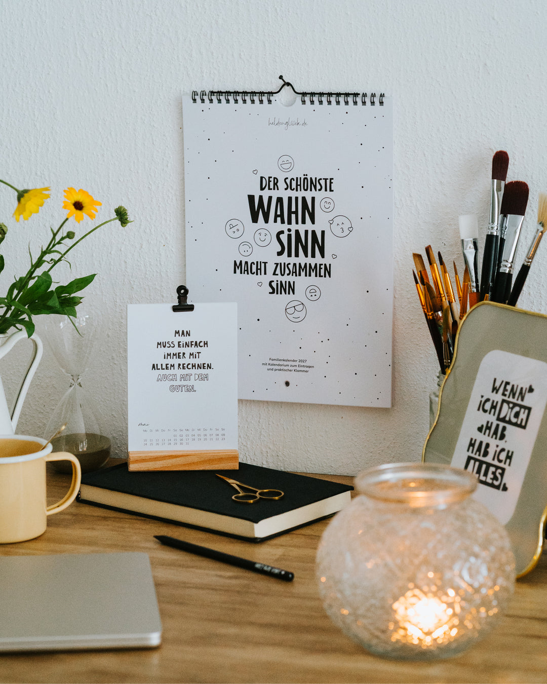 Kalender-Set 2027 im Homeoffice – Familienkalender an der Wand, Tischkalender auf dem Schreibtisch, daneben Windlicht.