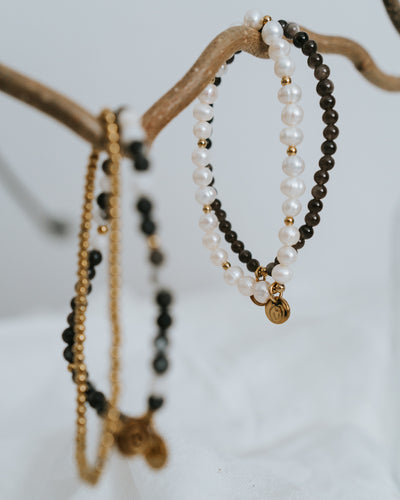 Süßwasserperlen-Armband in Gold neben Obsidian-Armband arrangiert.