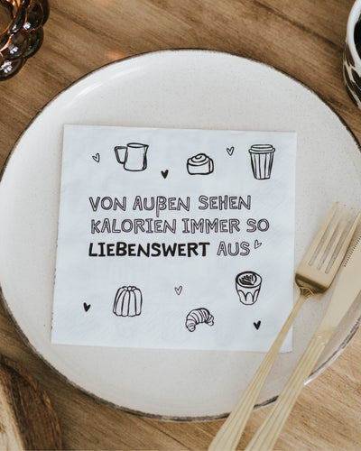 Serviette mit Spruch „Von außen sehen Kalorien immer so liebenswert aus“ auf gedecktem Tisch.