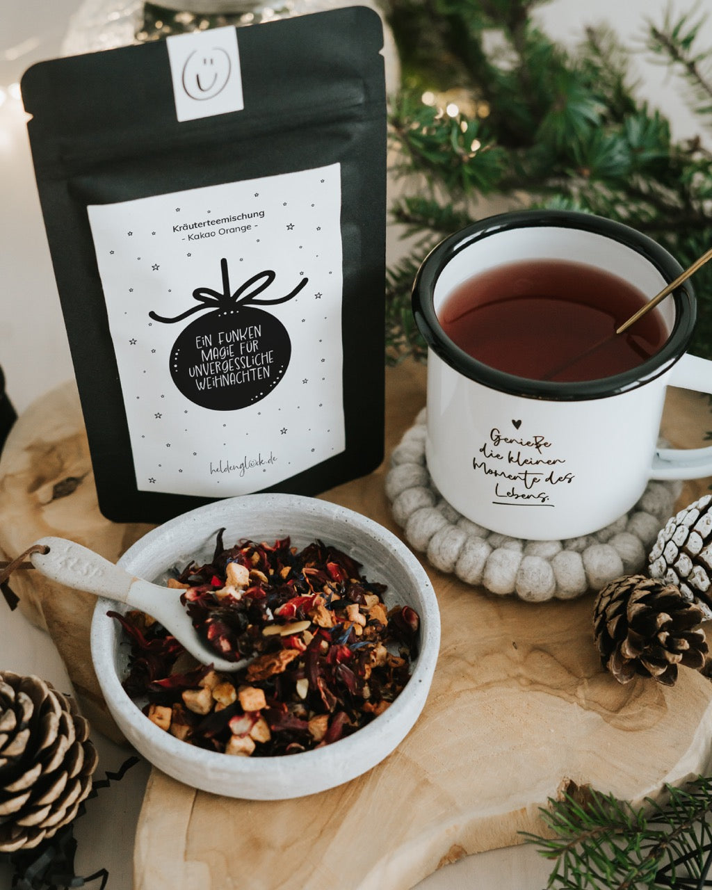 Weihnachtstee „Ein Funken Magie“ neben Becher mit aufgebrühtem Tee und Schale mit losem Tee – Tasse und Deko nicht enthalten.
