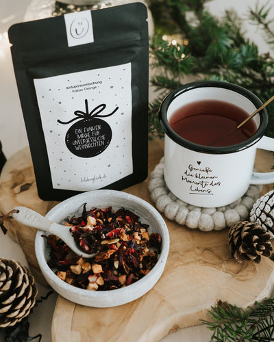 Weihnachtstee „Ein Funken Magie“ neben Becher mit aufgebrühtem Tee und Schale mit losem Tee – Tasse und Deko nicht enthalten.