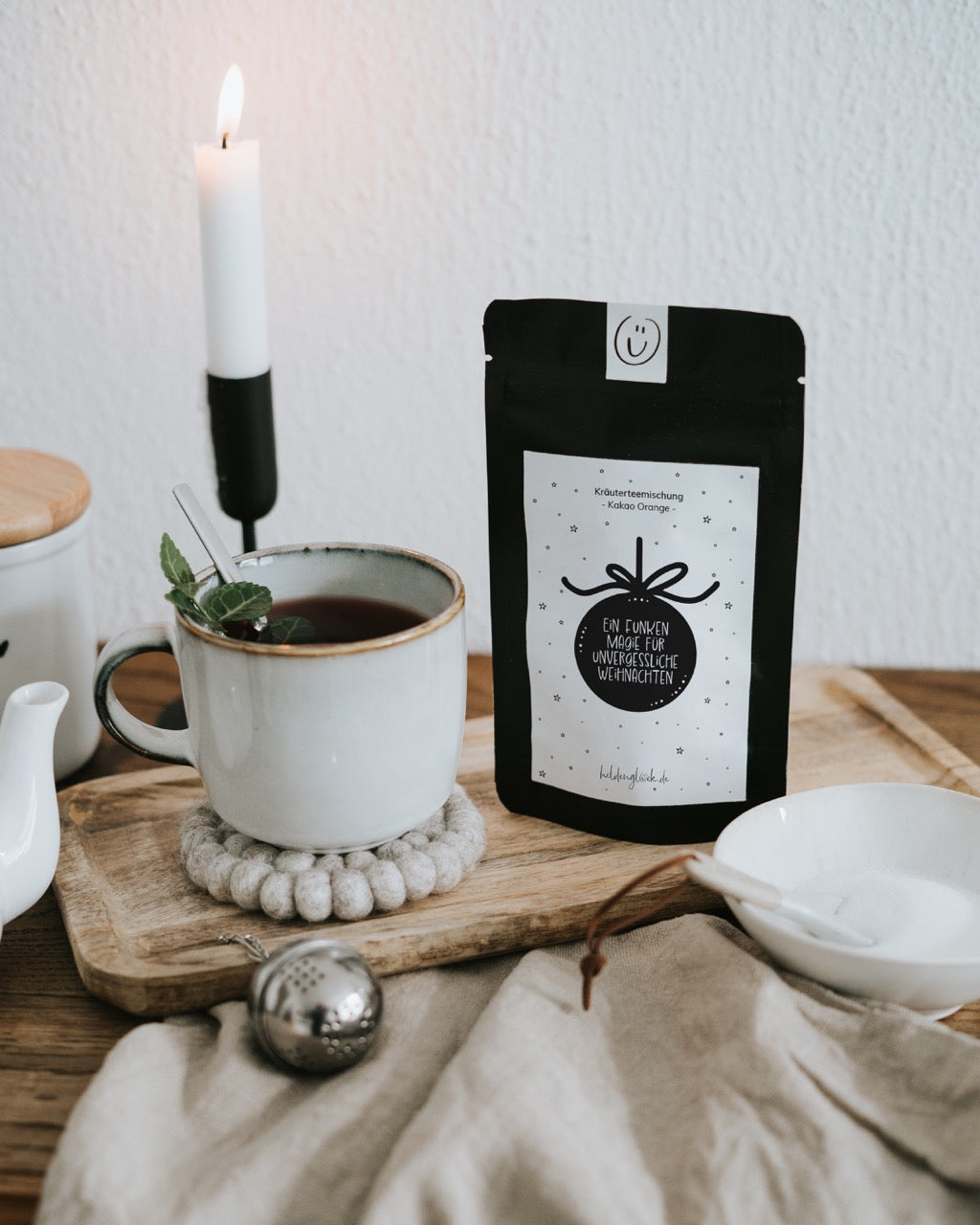 Weihnachtstee „Ein Funken Magie“ neben Tasse und Kerze – 35 g loser Tee, in Deutschland abgefüllt.