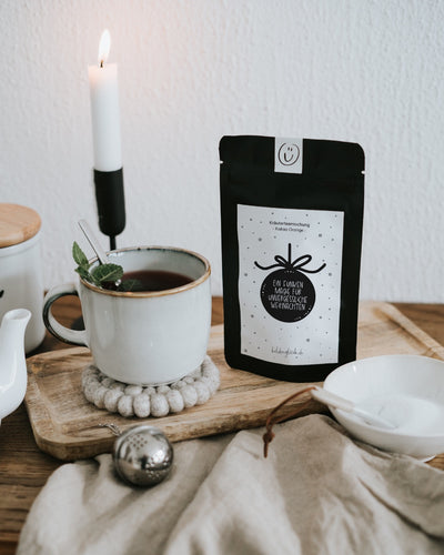 Weihnachtstee „Ein Funken Magie“ neben Tasse und Kerze – 35 g loser Tee, in Deutschland abgefüllt.