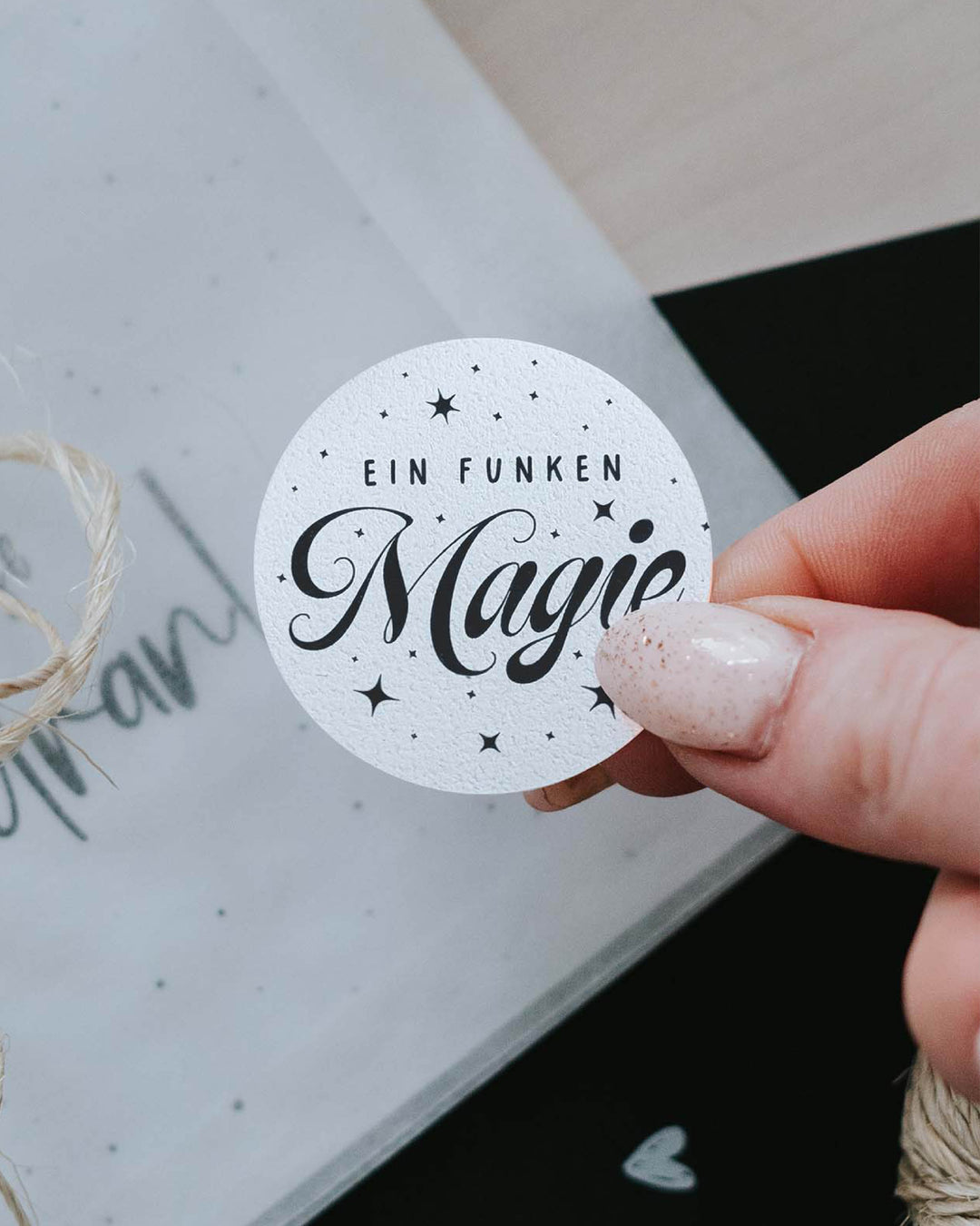 Hand klebt runden Aufkleber „Ein Funken Magie“ auf ein in Transparentpapier gewickeltes Weihnachtsgeschenk mit Kordel