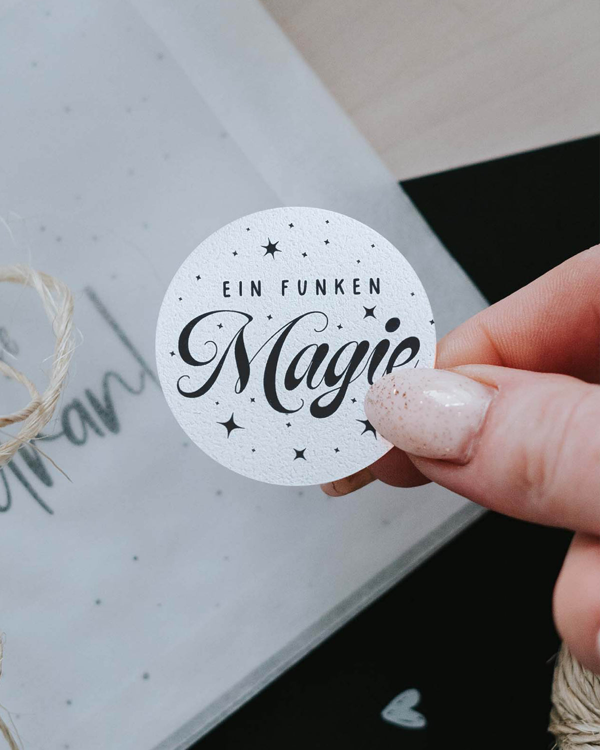 Hand klebt runden Aufkleber „Ein Funken Magie“ auf ein in Transparentpapier gewickeltes Weihnachtsgeschenk mit Kordel
