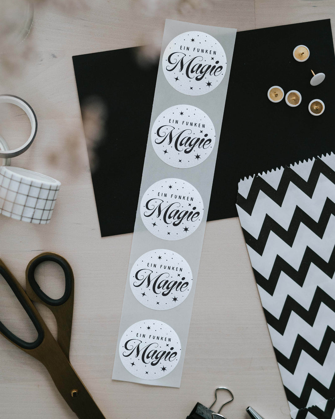 Streifen mit 5 Aufklebern „Ein Funken Magie“ neben Schere, Washi-Tape und gestreifter Geschenktüte