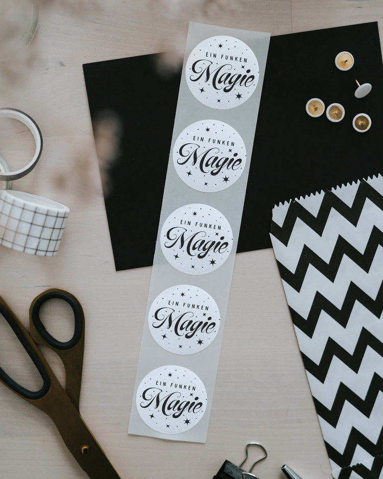 Streifen mit 5 Aufklebern „Ein Funken Magie“ neben Schere, Washi-Tape und gestreifter Geschenktüte