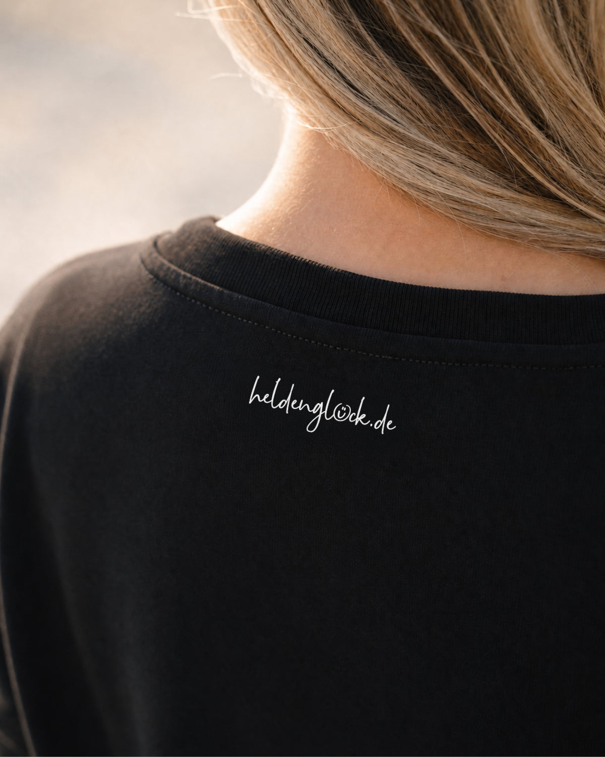 Weißer Schriftzug „heldenglück.de“ außen im Nacken des schwarzen T-Shirts, direkt unter dem Kragen.