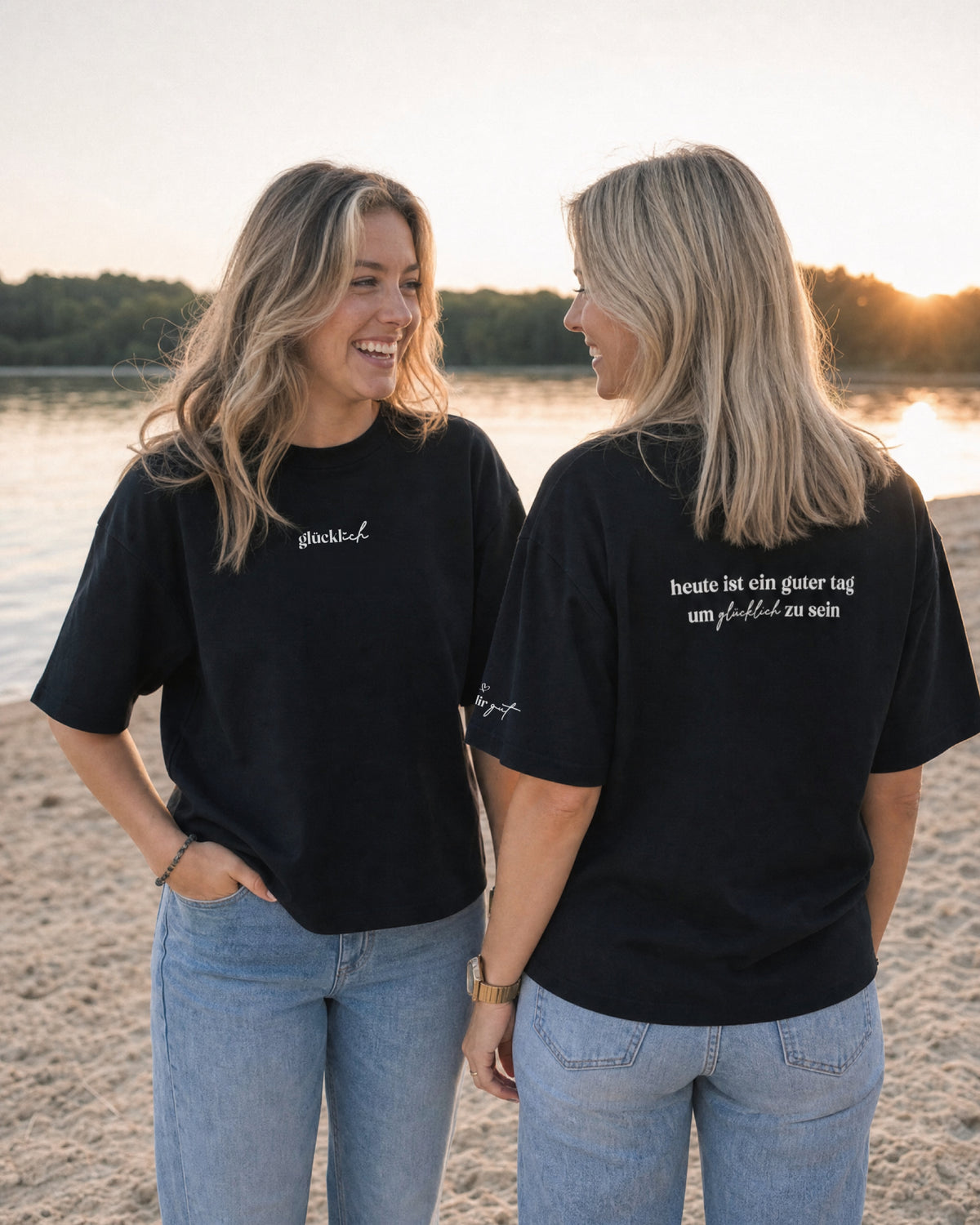 Zwei Frauen in schwarzen T-Shirts am Wasser; eine zeigt den Rückenprint „heute ist ein guter tag um glücklich zu sein“.