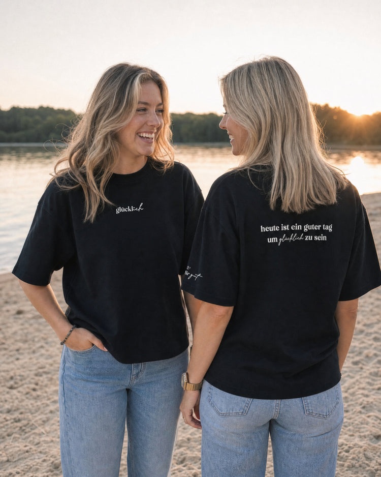 Zwei Frauen in schwarzen T-Shirts am Wasser; eine zeigt den Rückenprint „heute ist ein guter tag um glücklich zu sein“.