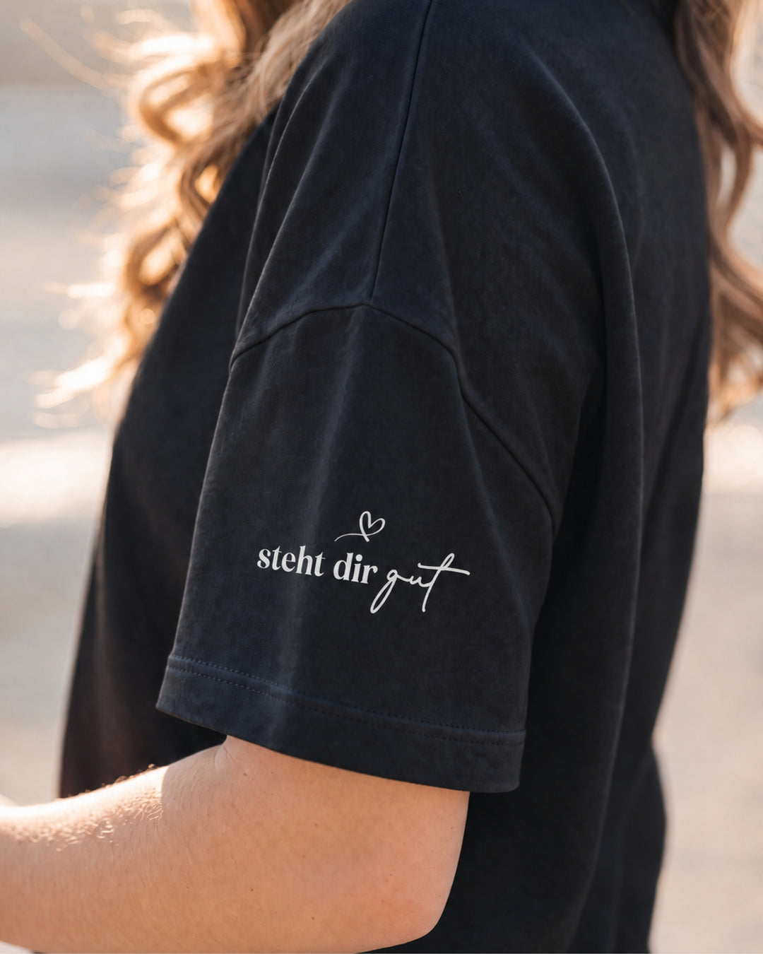Nahaufnahme des schwarzen T-Shirt-Ärmels mit weißem Herz und dem Schriftzug „steht dir gut“.