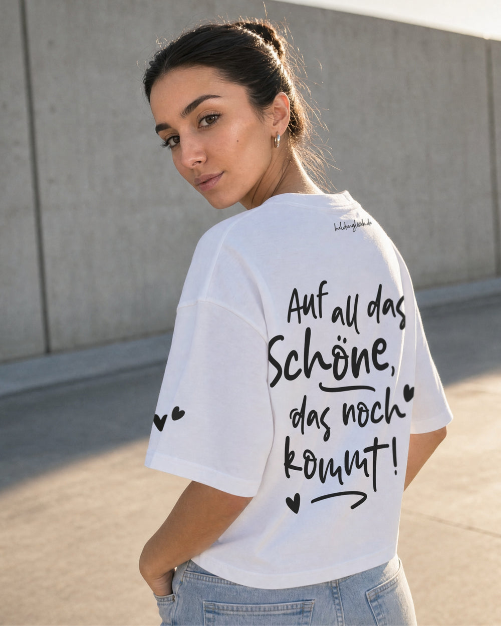 Frau im weißen T-Shirt von schräg hinten; Rückenprint „Auf all das Schöne, das noch kommt!“ mit Herzen und Pfeil.