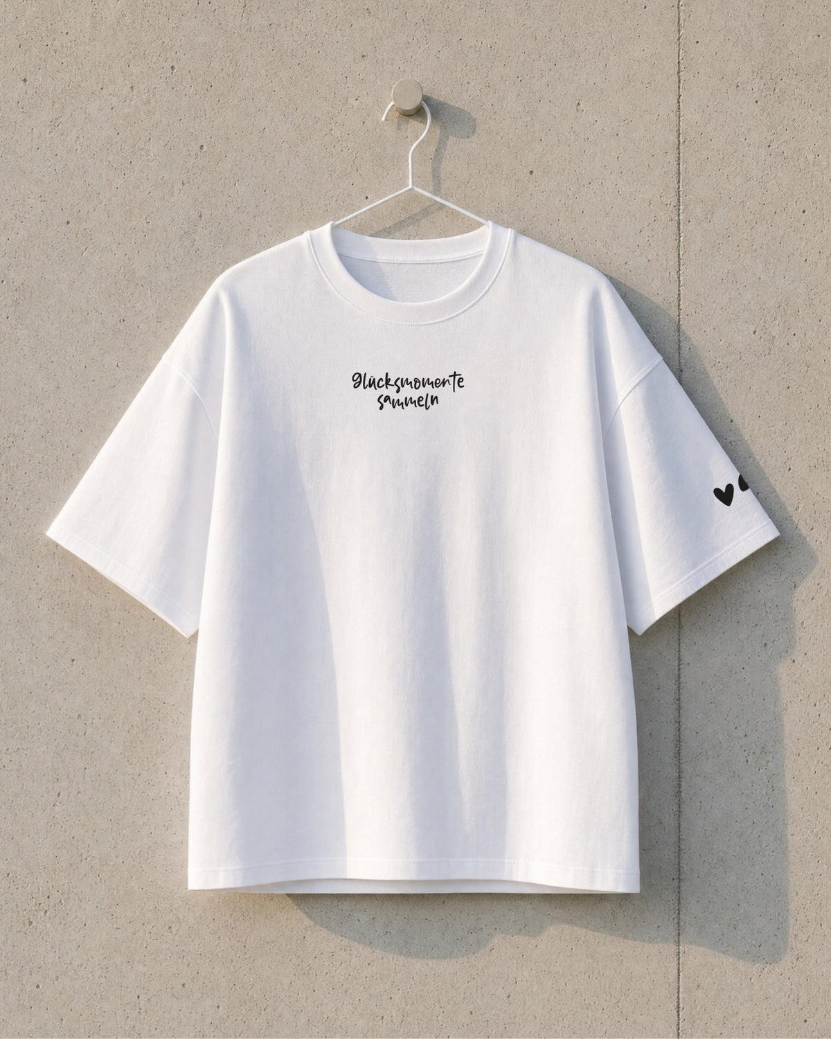 Vorderseite des weißen T-Shirts am Bügel, mit kleinem Schriftzug „Glücksmomente sammeln“ und zwei Herzen am Ärmel.