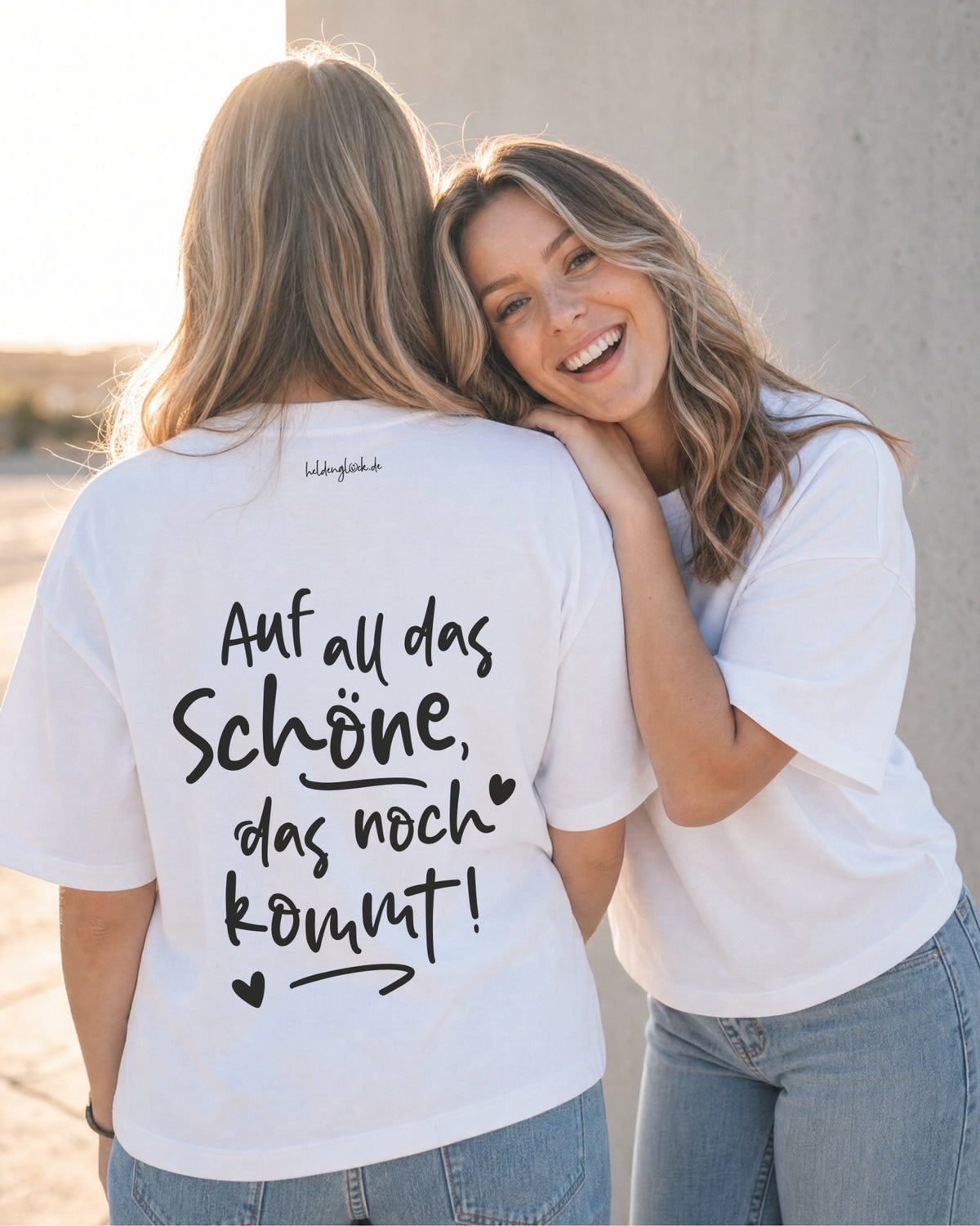 Zwei Frauen in weißen T-Shirts; eine zeigt den Rückenprint „Auf all das Schöne, das noch kommt!“ mit Herzen und Pfeil.