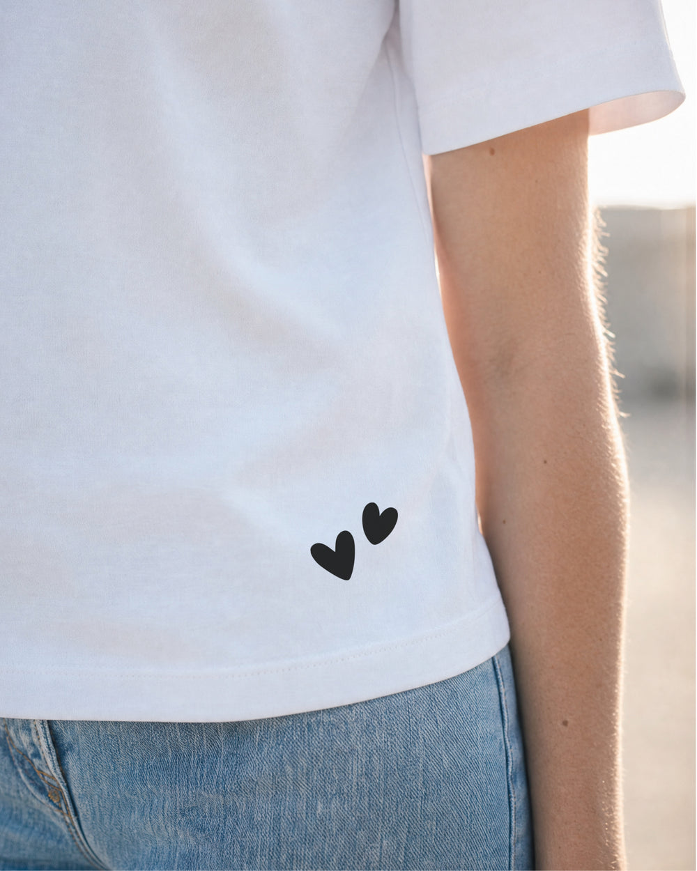Zwei kleine schwarze Herzen auf der Vorderseite des weißen T-Shirts, unten nahe dem Saum.