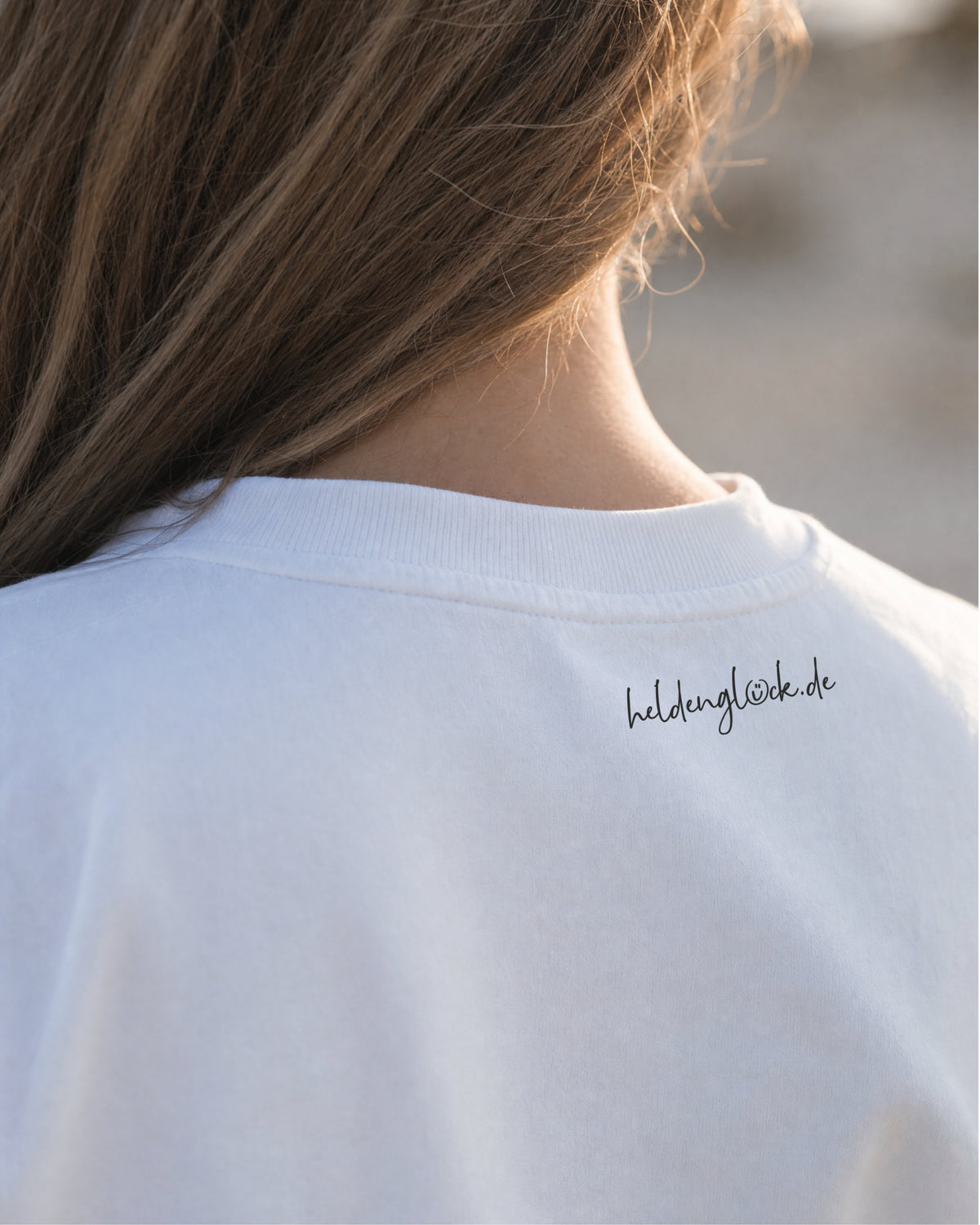Schwarzer Schriftzug „heldenglück.de“ außen im Nacken des weißen T-Shirts, direkt unter dem Kragen.