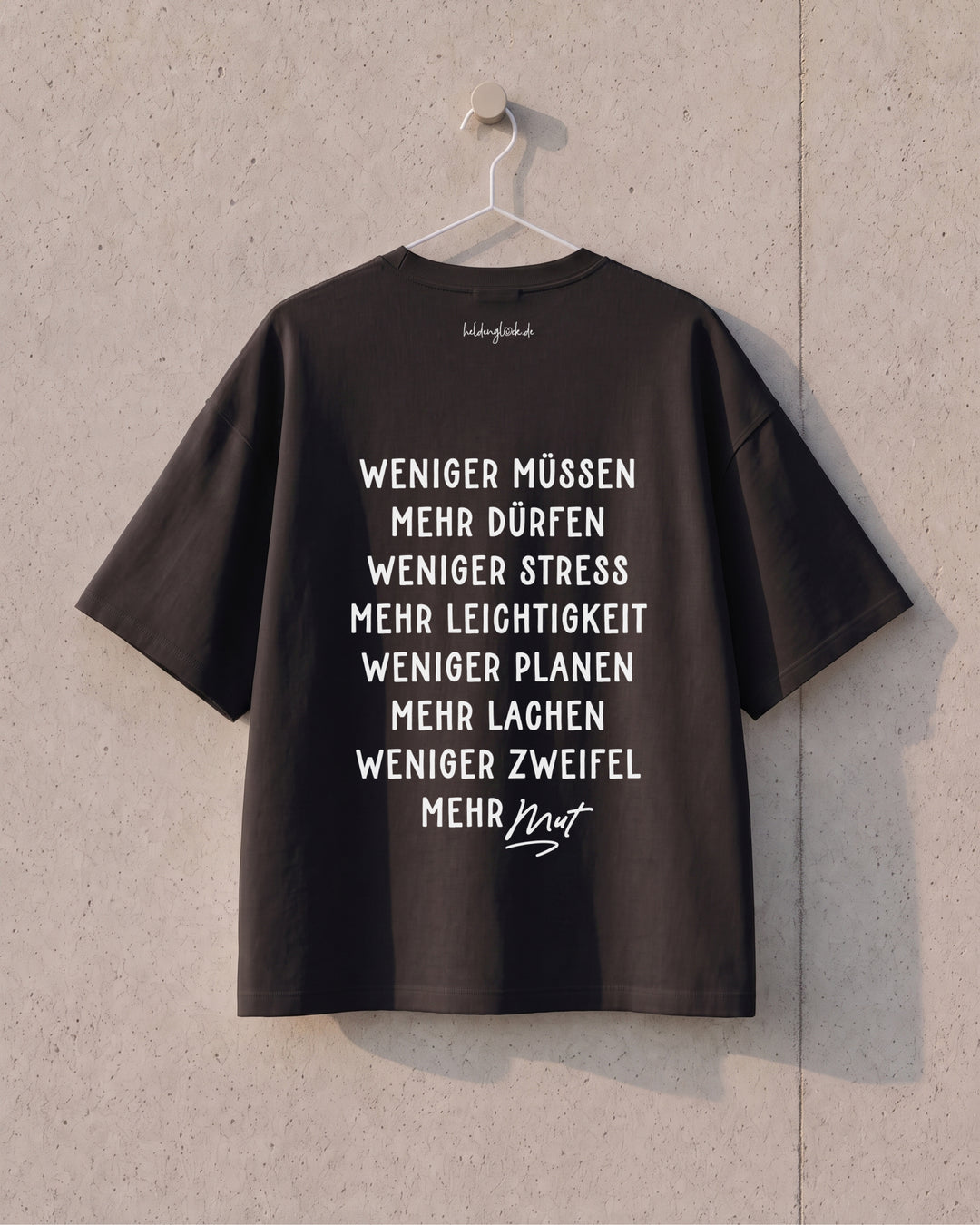 Schwarzes T-Shirt am Bügel mit den Rückenbotschaften „Weniger müssen, mehr dürfen“ bis „Weniger Zweifel, mehr Mut“.