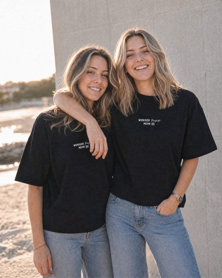 Zwei Frauen nebeneinander im schwarzen T-Shirt mit dem kleinen Brustaufdruck „Weniger perfekt, mehr du“.