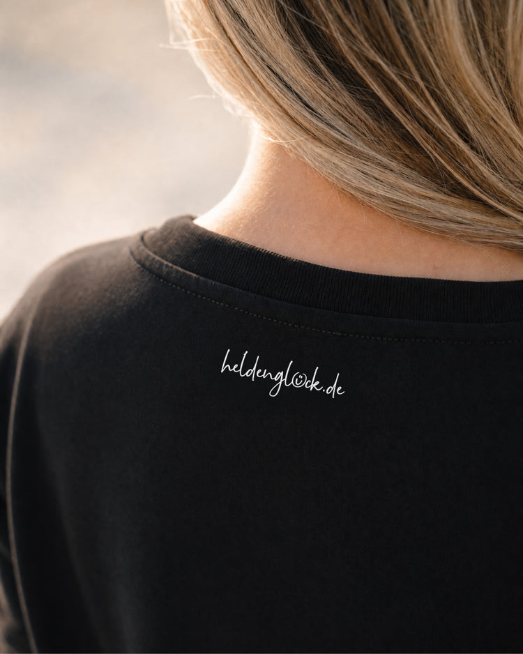 Weißer Schriftzug „heldenglück.de“ außen im Nacken des schwarzen T-Shirts, direkt unter dem Kragen.