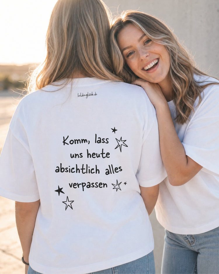 Zwei Frauen in weißen T-Shirts; auf einem Rücken steht „Komm, lass uns heute absichtlich alles verpassen“, umgeben von Sternen.