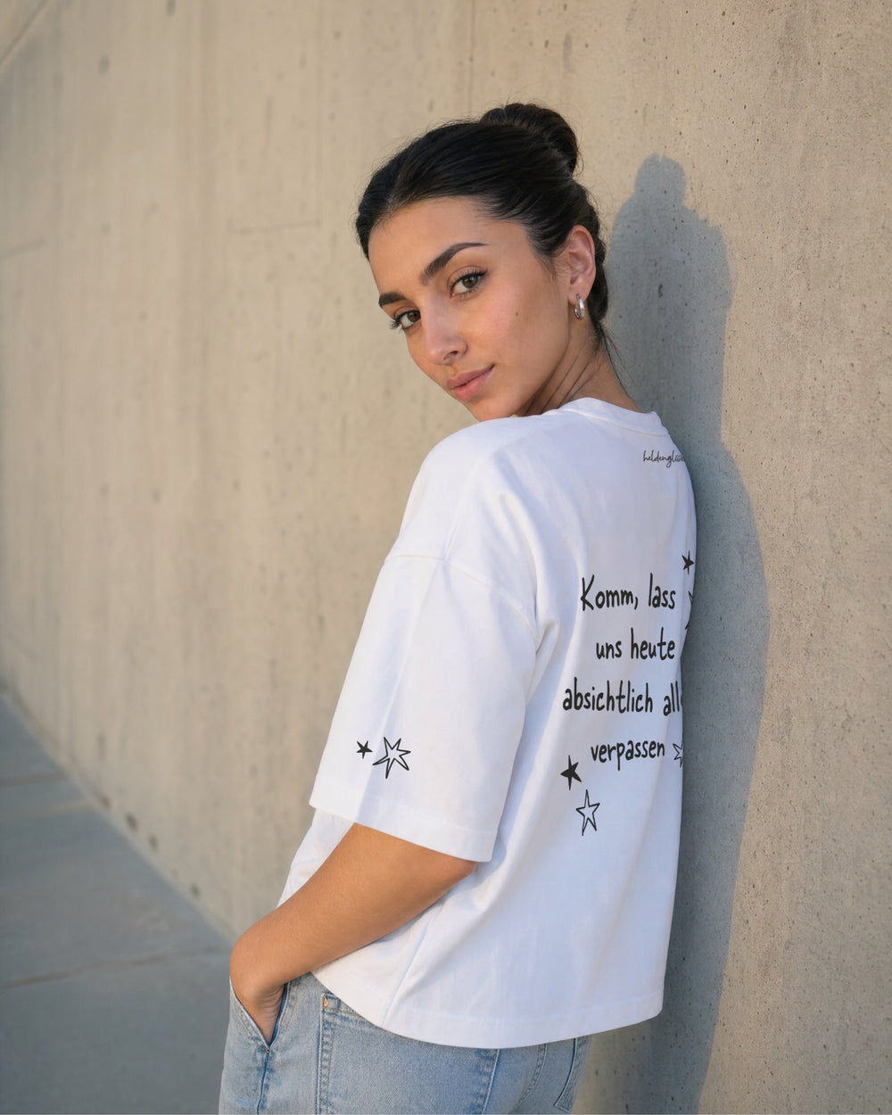 Weißes T-Shirt „Zeit verplempern“ an einer Frau von schräg hinten, mit großem Rückenprint und Sternen am Ärmel.