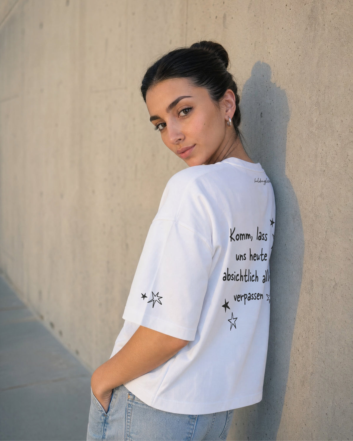 Weißes T-Shirt „Zeit verplempern“ an einer Frau von schräg hinten, mit großem Rückenprint und Sternen am Ärmel.