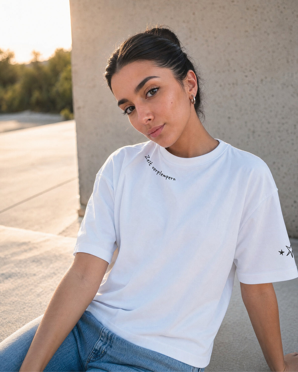 Vorderseite des weißen T-Shirts an einer sitzenden Frau, mit kleinem Schriftzug „Zeit verplempern“ und Sternen am Ärmel.