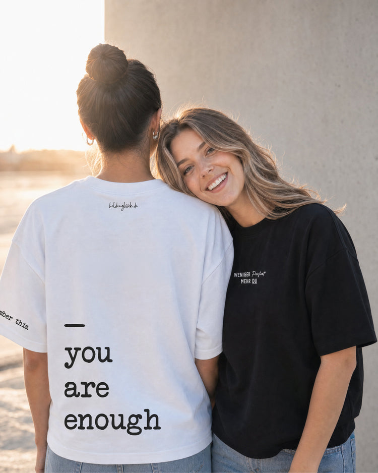 Zwei Frauen nebeneinander; eine zeigt das weiße T-Shirt mit Rückenprint „you are enough“, die andere trägt ein schwarzes Shirt.
